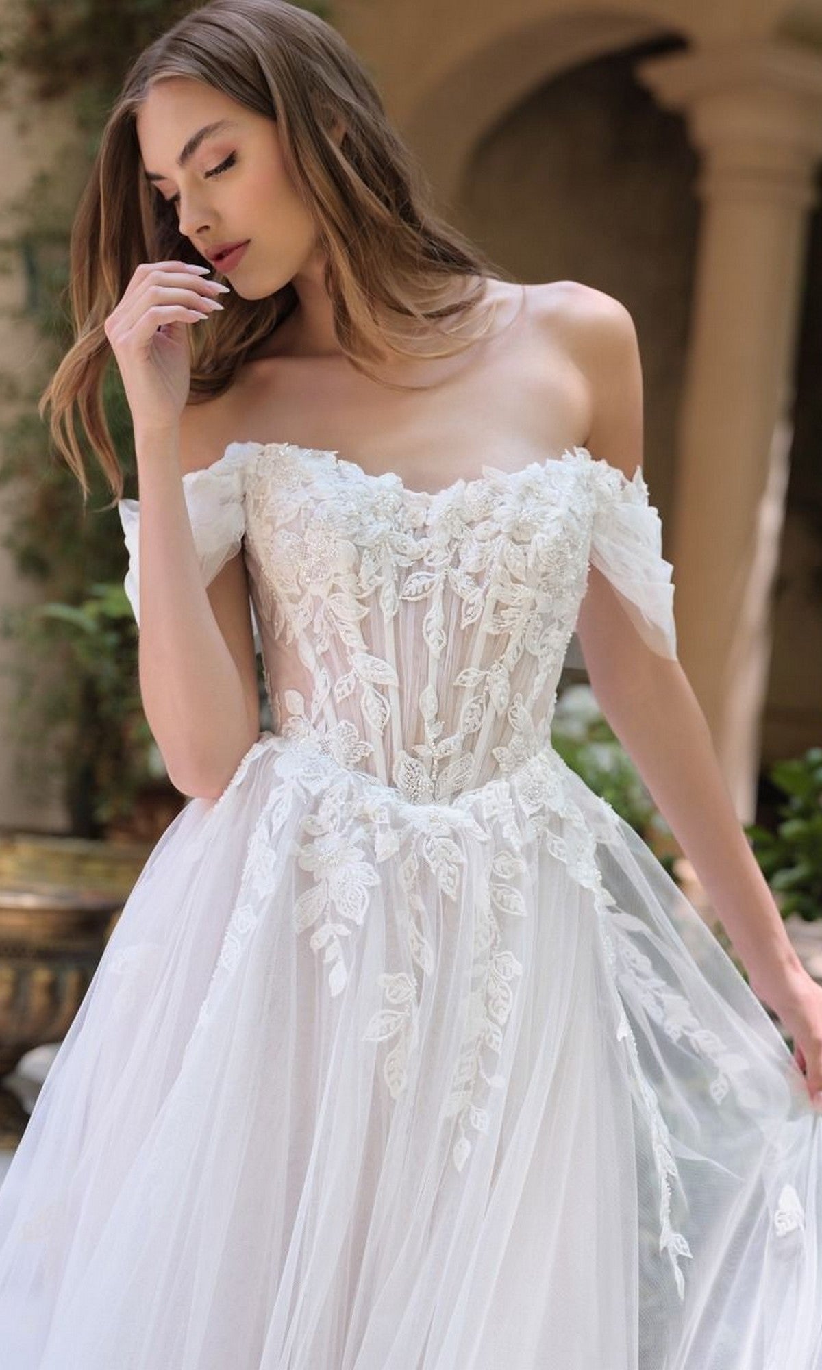 A woman in the Andrea and Leo WL089 long wedding dress, featuring off-the-shoulder floral lace appliques and an elegant A-line tulle skirt, stands gracefully in a charming courtyard setting.